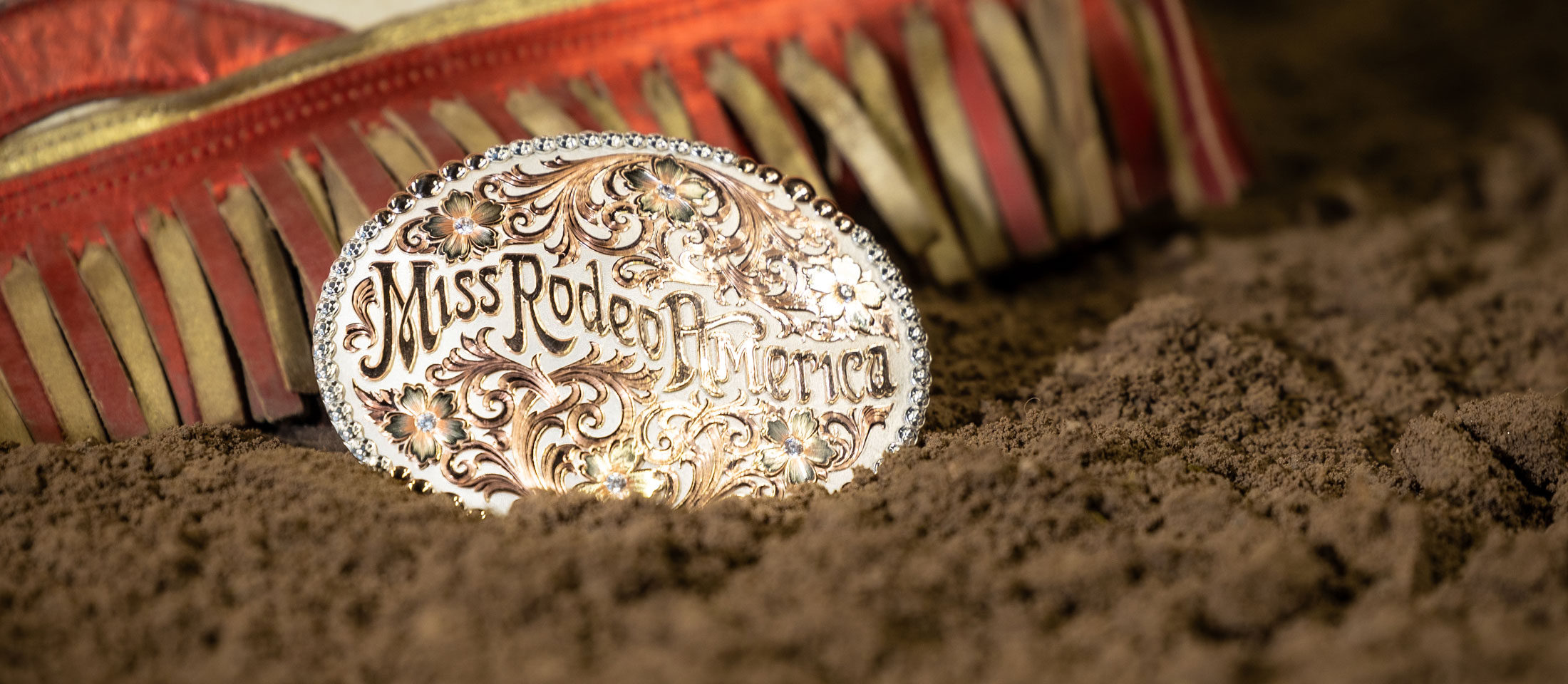 The silver and gold Miss Rodeo America buckle posed in arena dirt next to Miss Rodeo America chaps.